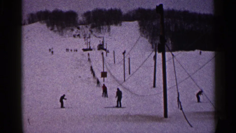 1960 Duluth home-movie frame showing skiers waiting on a snowy slope beside ski-lift poles and cables.