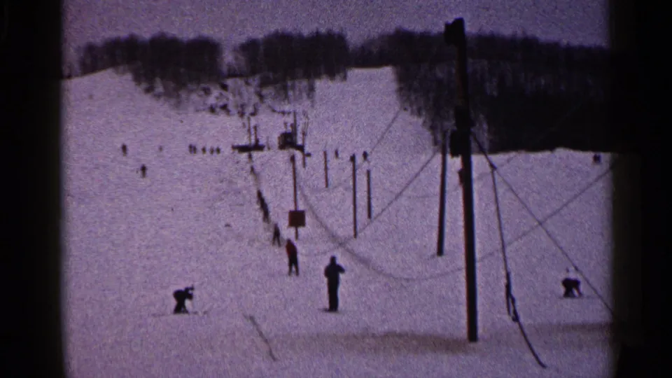 1960 Duluth frame with skiers spread across the snow near lift poles, cables, and a distant wooded ridge.