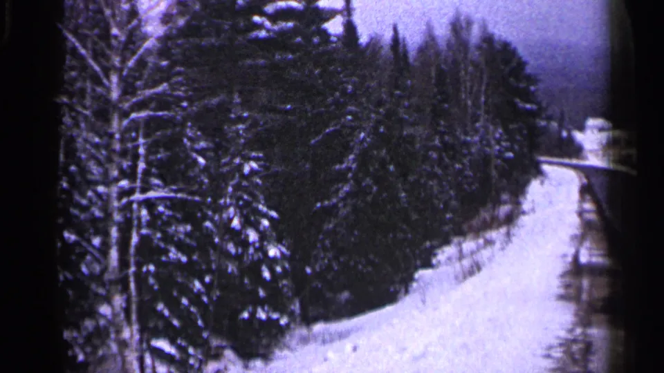1960 Duluth frame showing a snow-covered roadside or trail curving past dense winter trees, with distant pale structures unclear.
