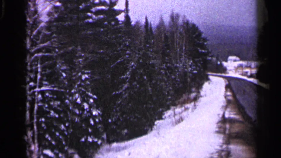 1960 Duluth home-movie frame of a curving snowy road or trail edged by forest, with indistinct buildings in the distance.
