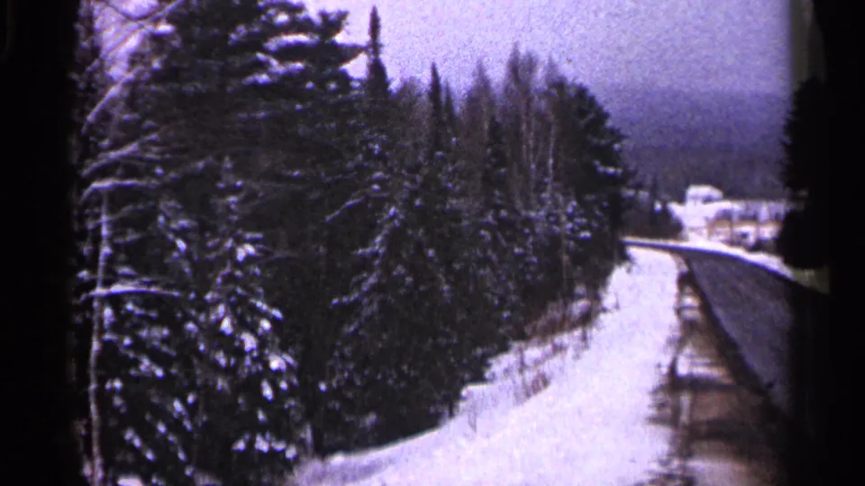 1960 Duluth-area 8mm frame showing a snow-lined winter road bordered by evergreens, with a distant structure softened by haze.