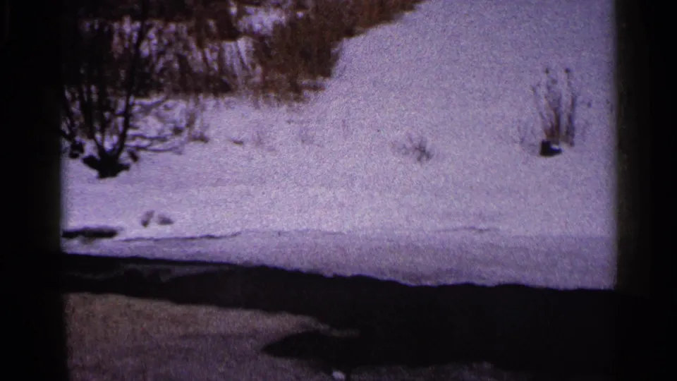 1960 Duluth-area 8mm frame of a snowy river or stream bank, with leafless brush and dark open water at the lower edge.