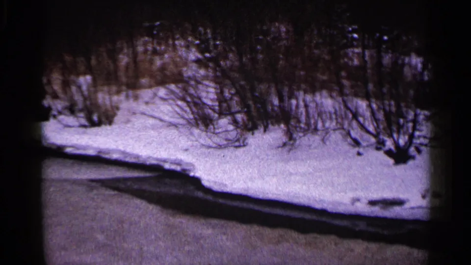 1960 Duluth winter frame with a partially frozen waterway, snow along the bank, and bare shrubs under a dark 8mm border.