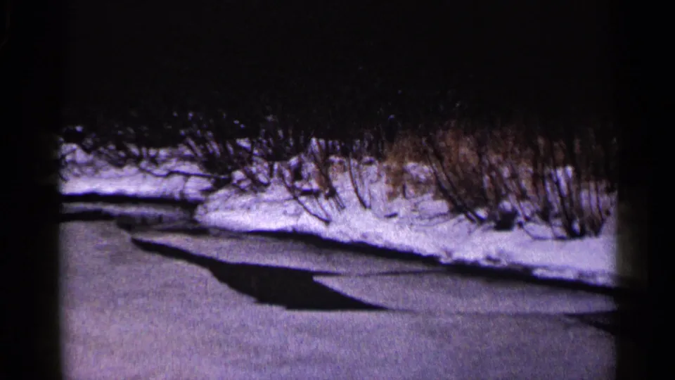 1960 Duluth frame showing a darker break in the frozen stream surface near a snow-covered bank and winter shrubs.