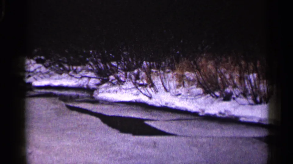 1960 Duluth home-movie frame showing the frozen stream surface, a dark ice edge, snow bank, and leafless shrubs along the waterway.