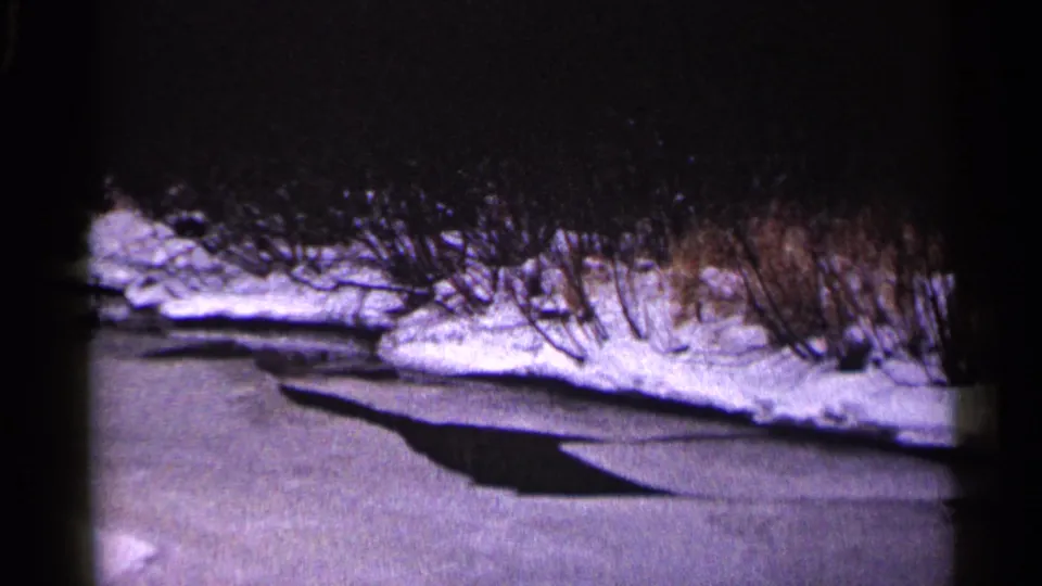 1960 Duluth home-movie frame showing a frozen waterway, snowy bank, bare brush, and a long dark shadow across the ice.