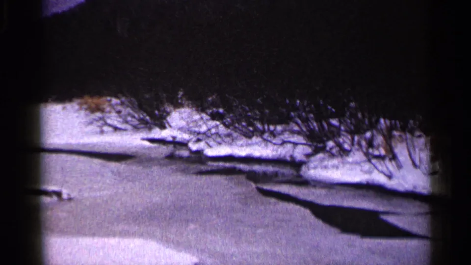 1960 Duluth 8mm frame showing a frozen water edge, snow patches, bare winter brush, and a dark shadow crossing the scene.