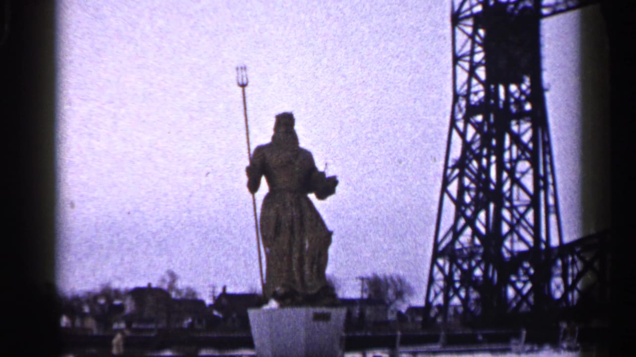 Wider 1960 Duluth view of the Neptune statue with trident, cloudy sky, tower, and distant buildings.
