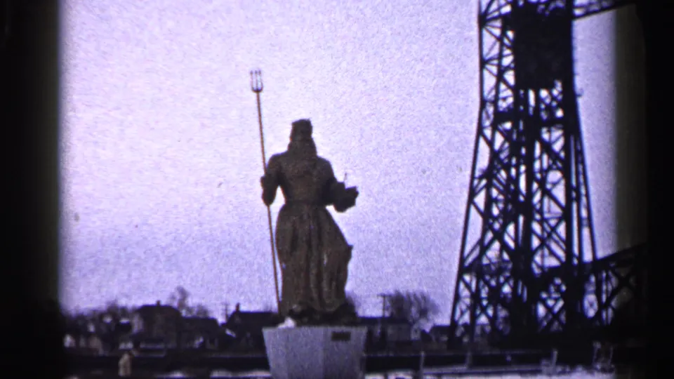 Wider 1960 Duluth view of the Neptune statue with trident, cloudy sky, tower, and distant buildings.