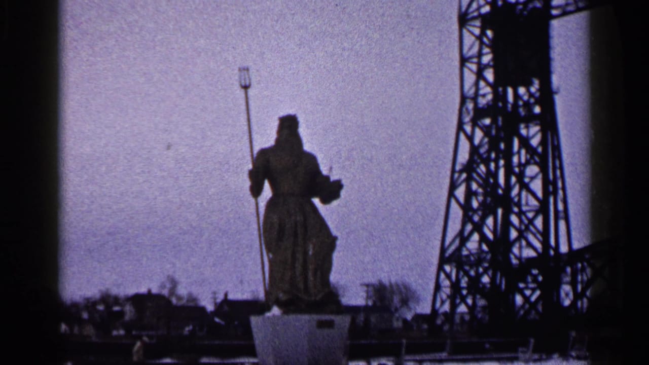1960 Duluth home-movie frame showing a robed trident-holding statue on a pedestal beside dark industrial bridge steel.