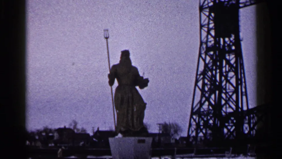 1960 Duluth home-movie frame showing a robed trident-holding statue on a pedestal beside dark industrial bridge steel.