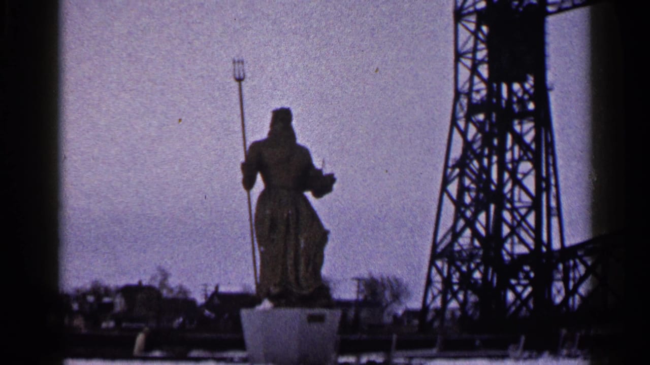 1960 Duluth 8mm frame holding on the robed trident statue, with industrial steelwork and waterfront details behind it.