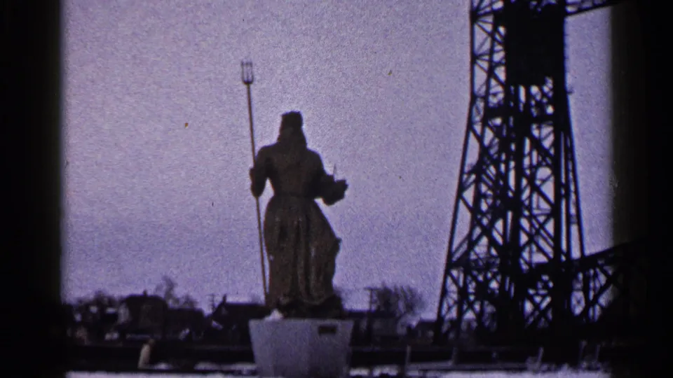 1960 Duluth 8mm frame holding on the robed trident statue, with industrial steelwork and waterfront details behind it.