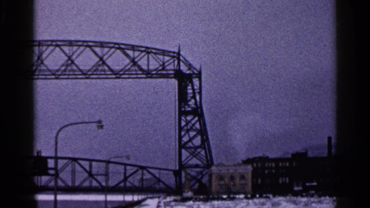 1960 Duluth frame shifting from the statue to a large industrial bridge span, streetlights, and waterfront buildings.