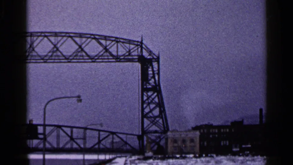 1960 Duluth frame shifting from the statue to a large industrial bridge span, streetlights, and waterfront buildings.
