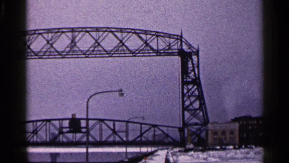 Duluth Aerial Lift Bridge in a 1960 winter 8mm view, with snowy ground, a lower span, streetlight, and pale sky.