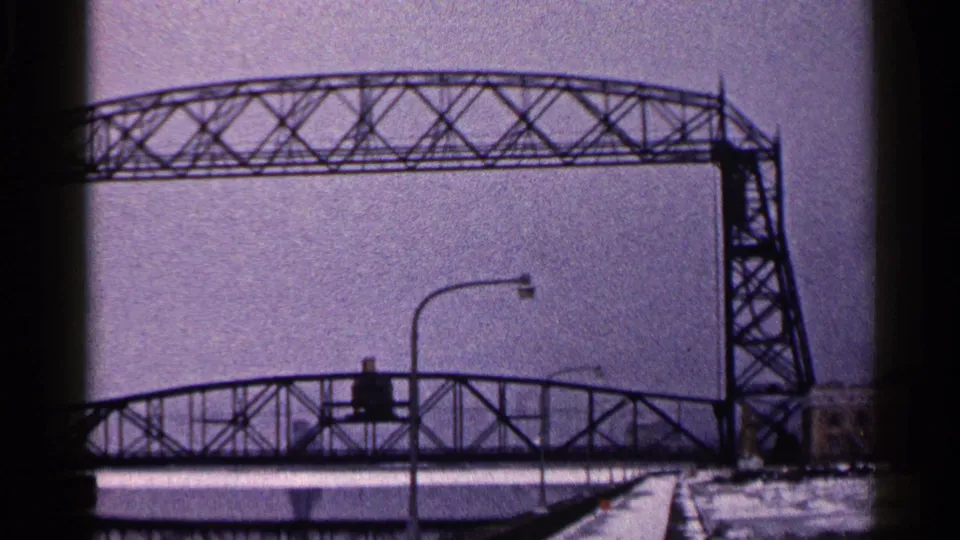 A 1960 Duluth winter frame shows the lift bridge truss crossing above a lower bridge span, with snow and a streetlight below.