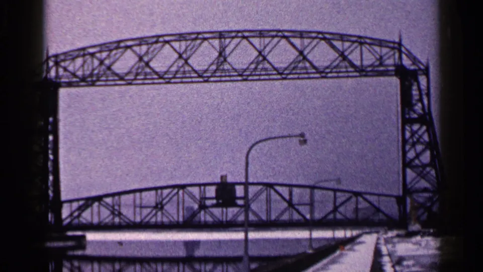1960 Duluth home-movie frame showing a large steel truss bridge with snowy foreground edges and curved streetlights.