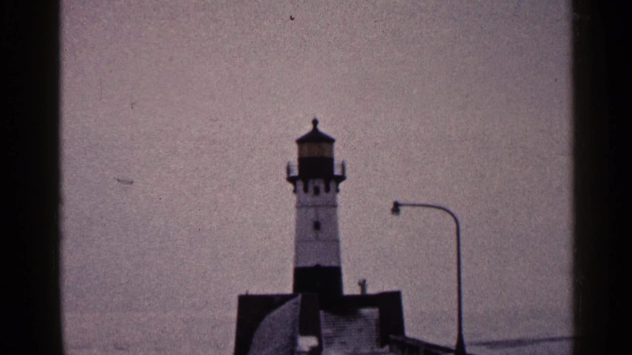 1960 Duluth 8mm frame with the lighthouse centered beyond dark vertical borders, a curved lamppost at right, and muted sky behind it.