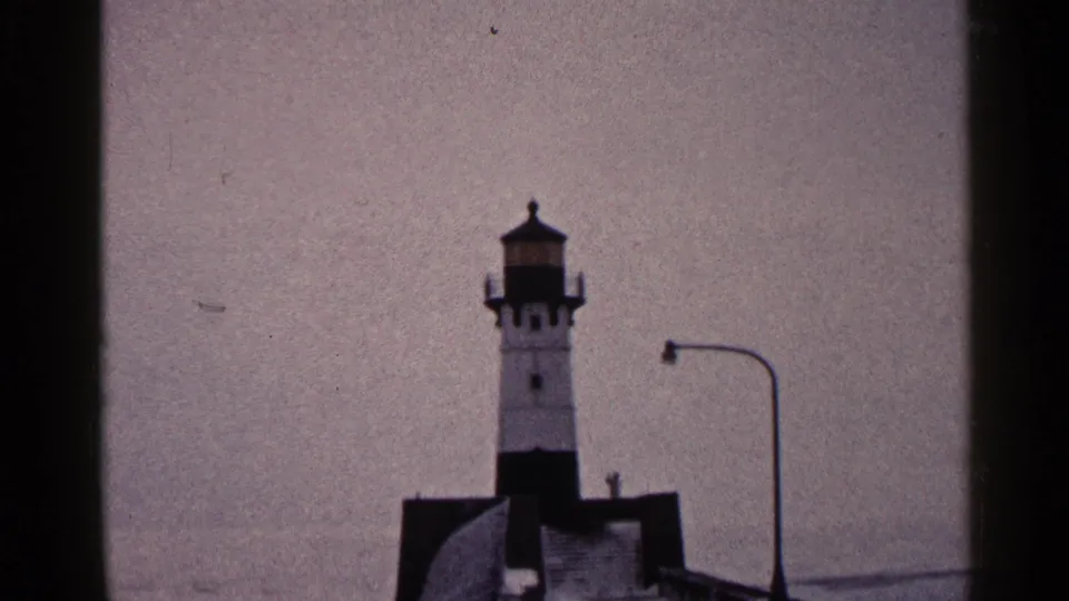 1960 Duluth 8mm frame with the lighthouse centered beyond dark vertical borders, a curved lamppost at right, and muted sky behind it.