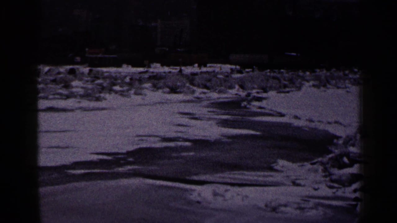 1960 Duluth frame showing icy shoreline and snow in front of a distant lighthouse, pier, lamppost, and low waterfront structures.