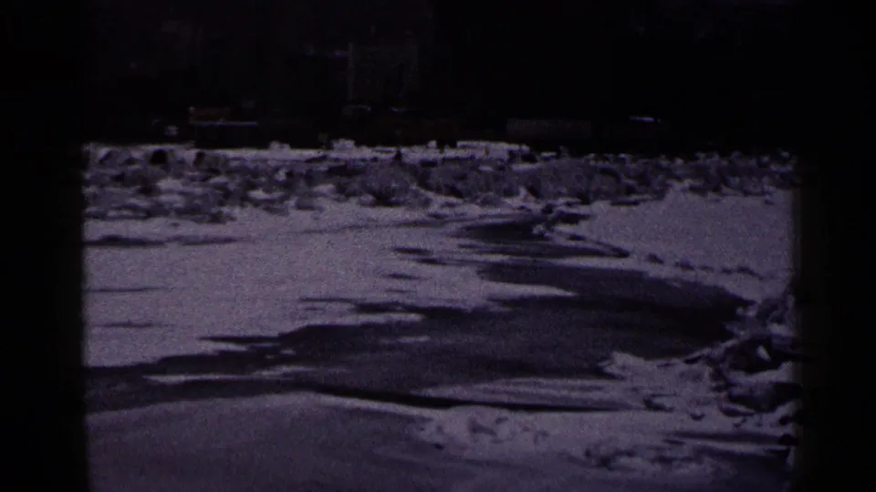 1960 Duluth frame showing icy shoreline and snow in front of a distant lighthouse, pier, lamppost, and low waterfront structures.