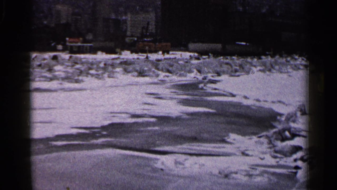 1960 Duluth home-movie frame with ice and snow across the foreground, a distant lighthouse, curved lamppost, and dark harbor background.
