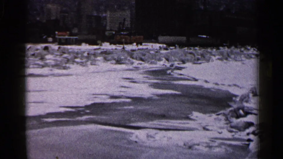 1960 Duluth home-movie frame with ice and snow across the foreground, a distant lighthouse, curved lamppost, and dark harbor background.