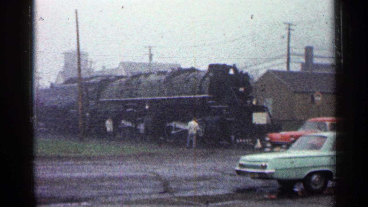 1960 Duluth home-movie frame showing a black steam locomotive near people and period cars under overcast light.