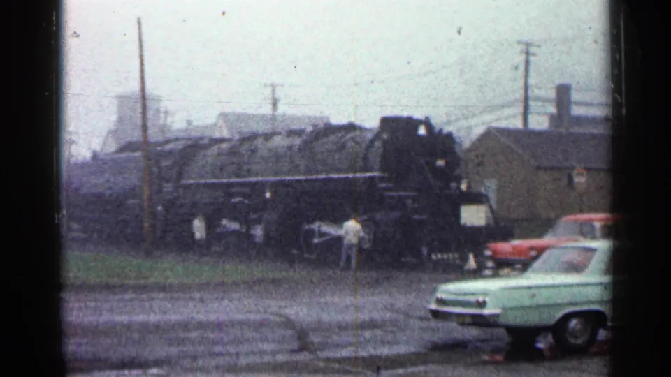 1960 Duluth home-movie frame showing a black steam locomotive near people and period cars under overcast light.