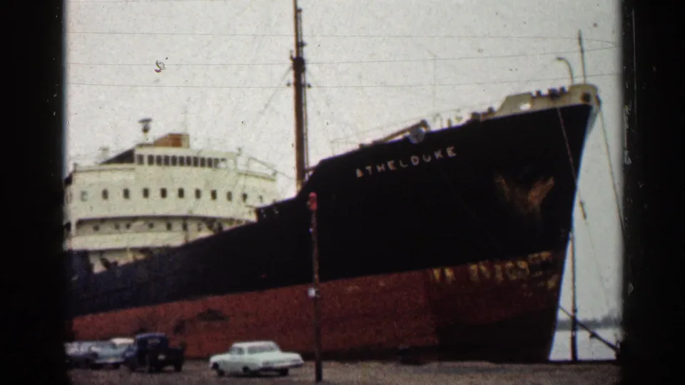 1960 Duluth frame showing the docked freighter ATHELDUKE, harbor poles, and mid-century cars at the waterfront.