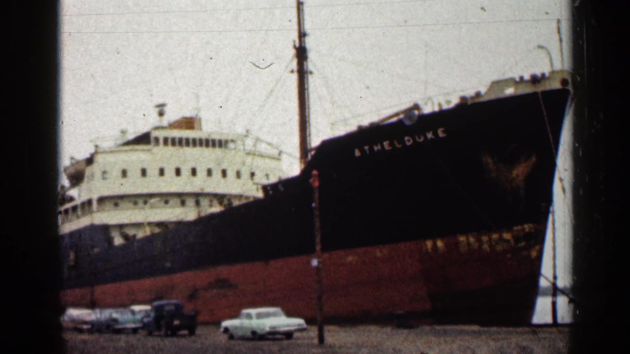 Large freighter at a dock with parked vintage cars in front.