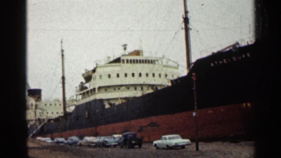 Duluth harbor frame showing the freighter side, white upper decks, a mast, dockside cars, and a dark pickup in the foreground.