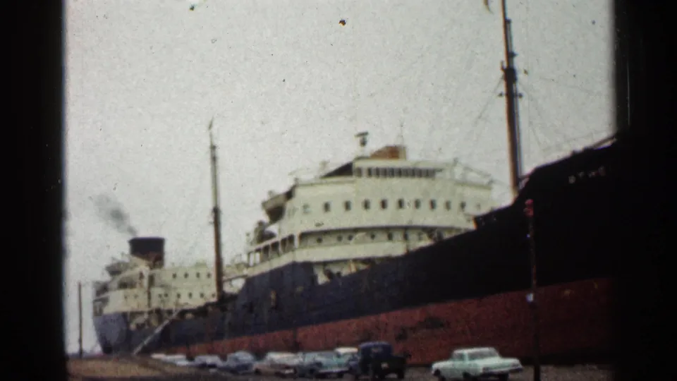 Athel Duke at the Duluth dock with smoke rising near the funnel, a red lower hull, white superstructure, and parked cars below.