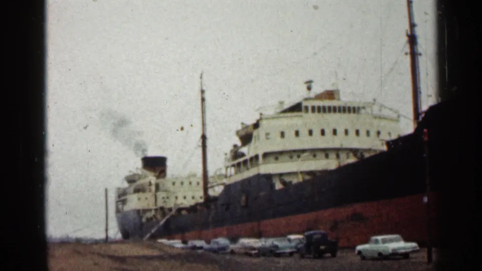 Large passenger ship at the Duluth waterfront in 1960, with a dark hull, white upper decks, smoke, and parked automobiles.