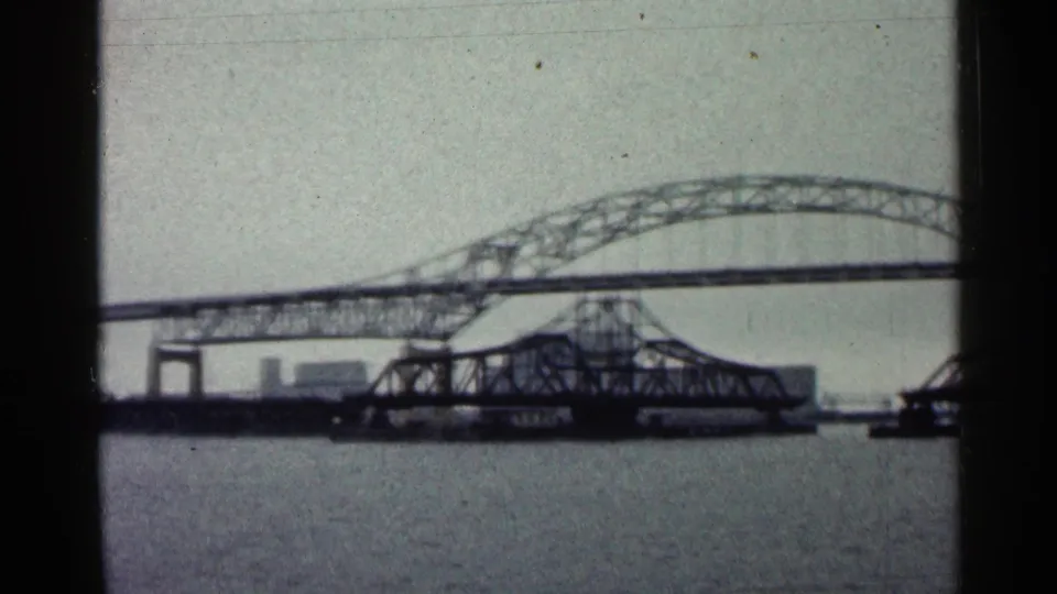 Final 1960 Duluth bridge frame showing arched steelwork, a lower truss section, river water, and distant shoreline structures.