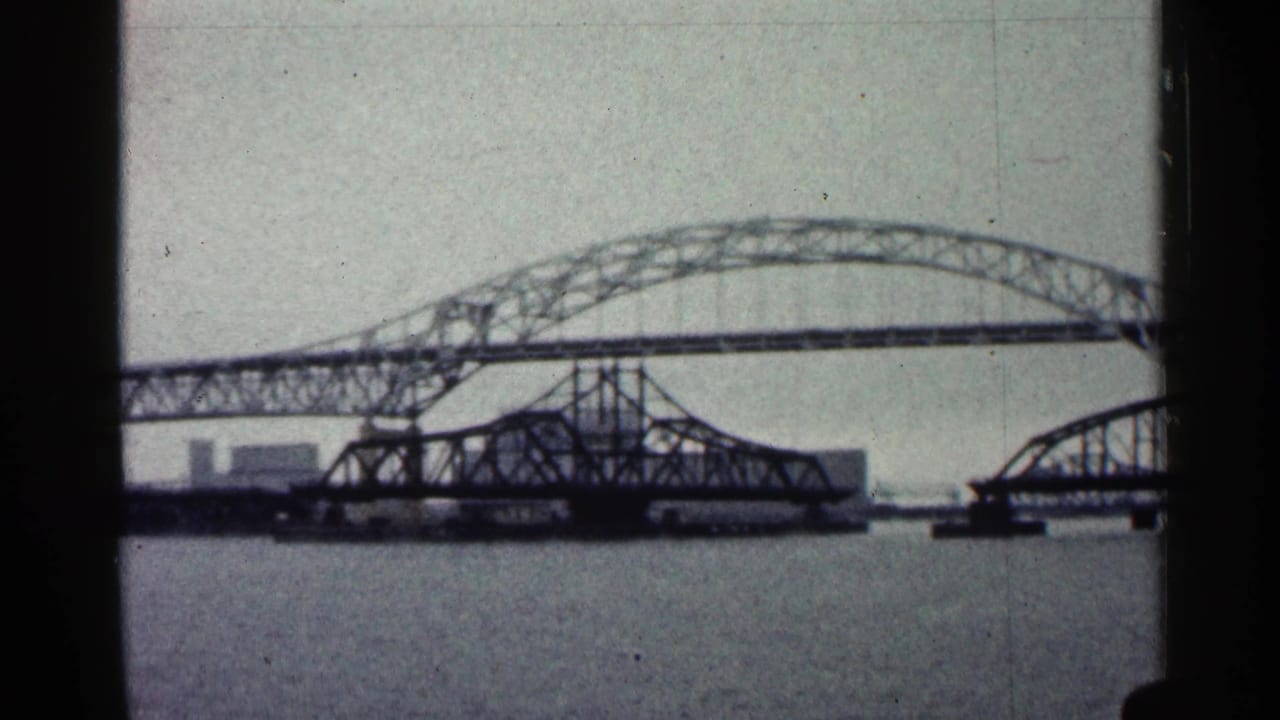 1960 Duluth waterfront view with a long arched steel bridge crossing a river and a lower bridge span beneath it.