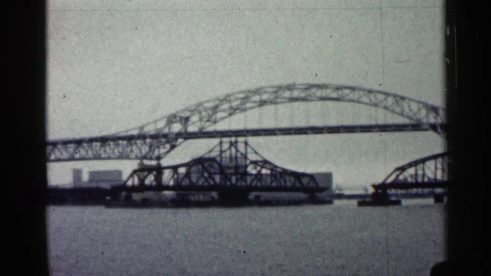 1960 Duluth waterfront view with a long arched steel bridge crossing a river and a lower bridge span beneath it.