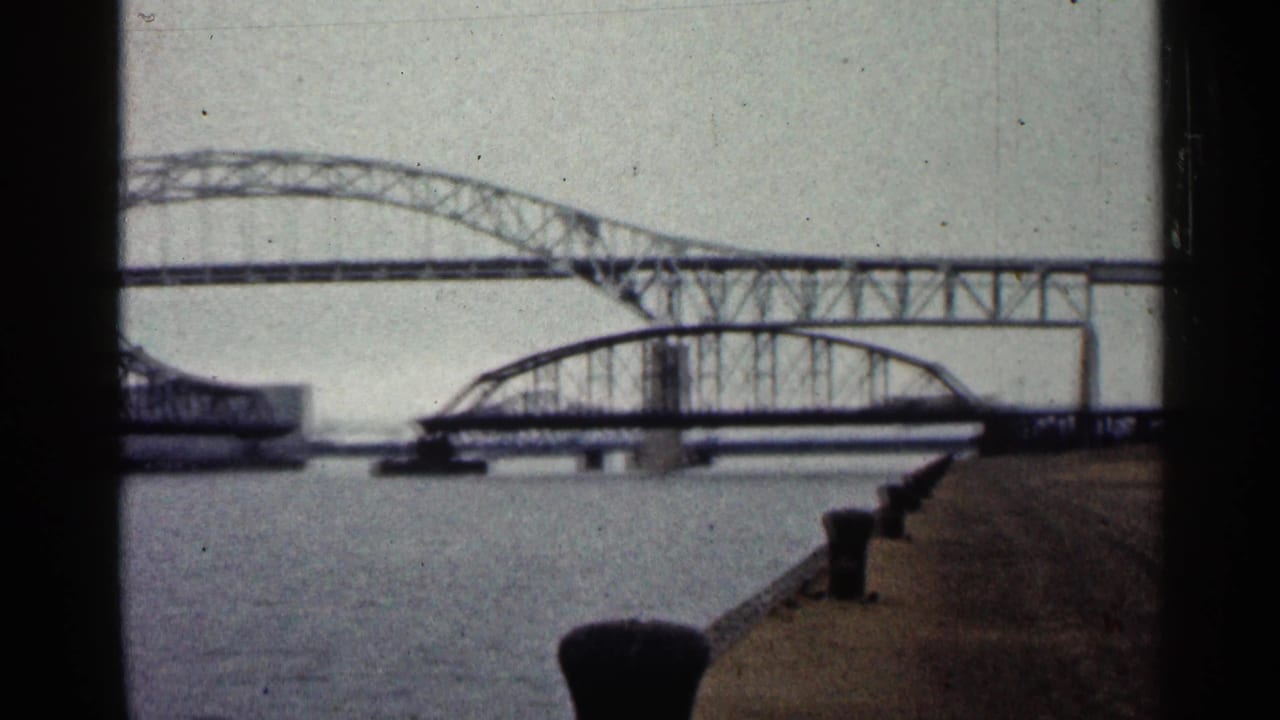 1960 Duluth waterfront perspective with bollards along a concrete pier and steel bridge spans crossing the river beyond.