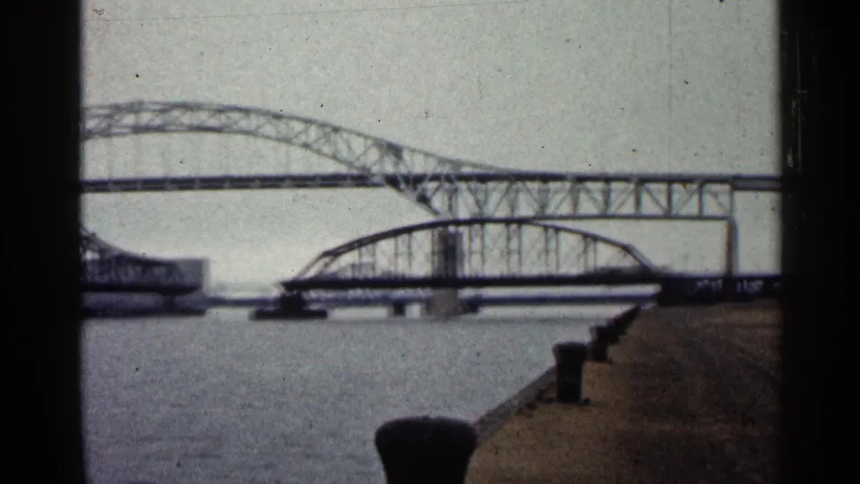 1960 Duluth waterfront perspective with bollards along a concrete pier and steel bridge spans crossing the river beyond.