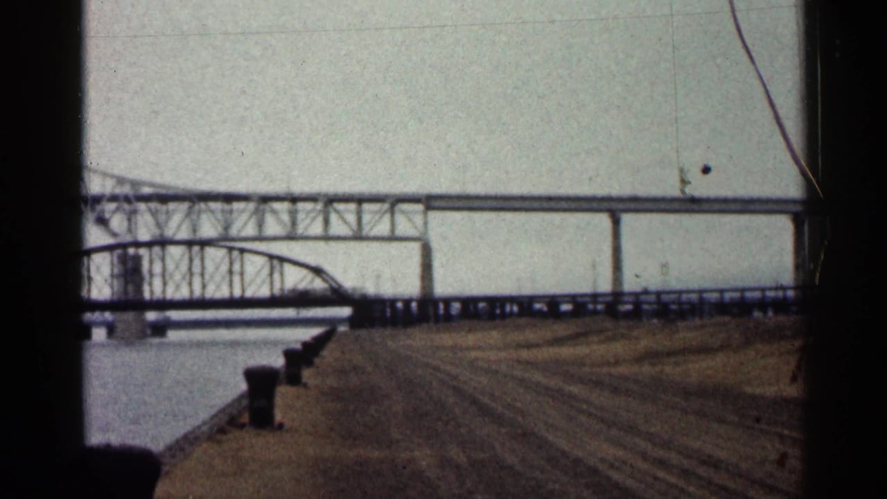 1960 Duluth home-movie frame showing a large bridge with a truss section, elevated span, water and bare foreground earth.