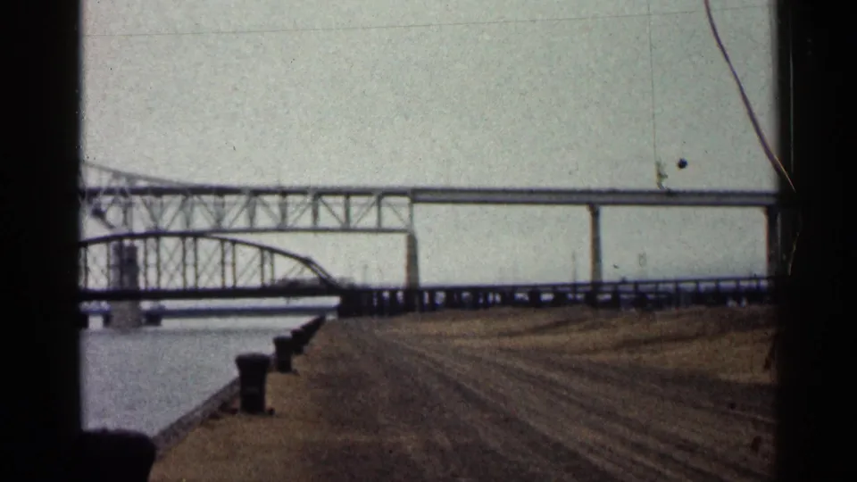 1960 Duluth home-movie frame showing a large bridge with a truss section, elevated span, water and bare foreground earth.