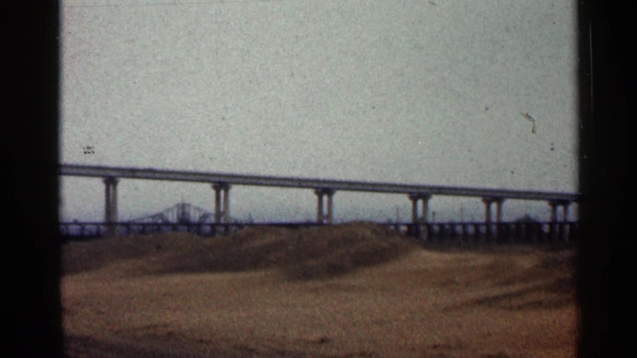 1960 Duluth frame showing elevated bridge supports, a distant truss section and open dirt foreground under a muted sky.