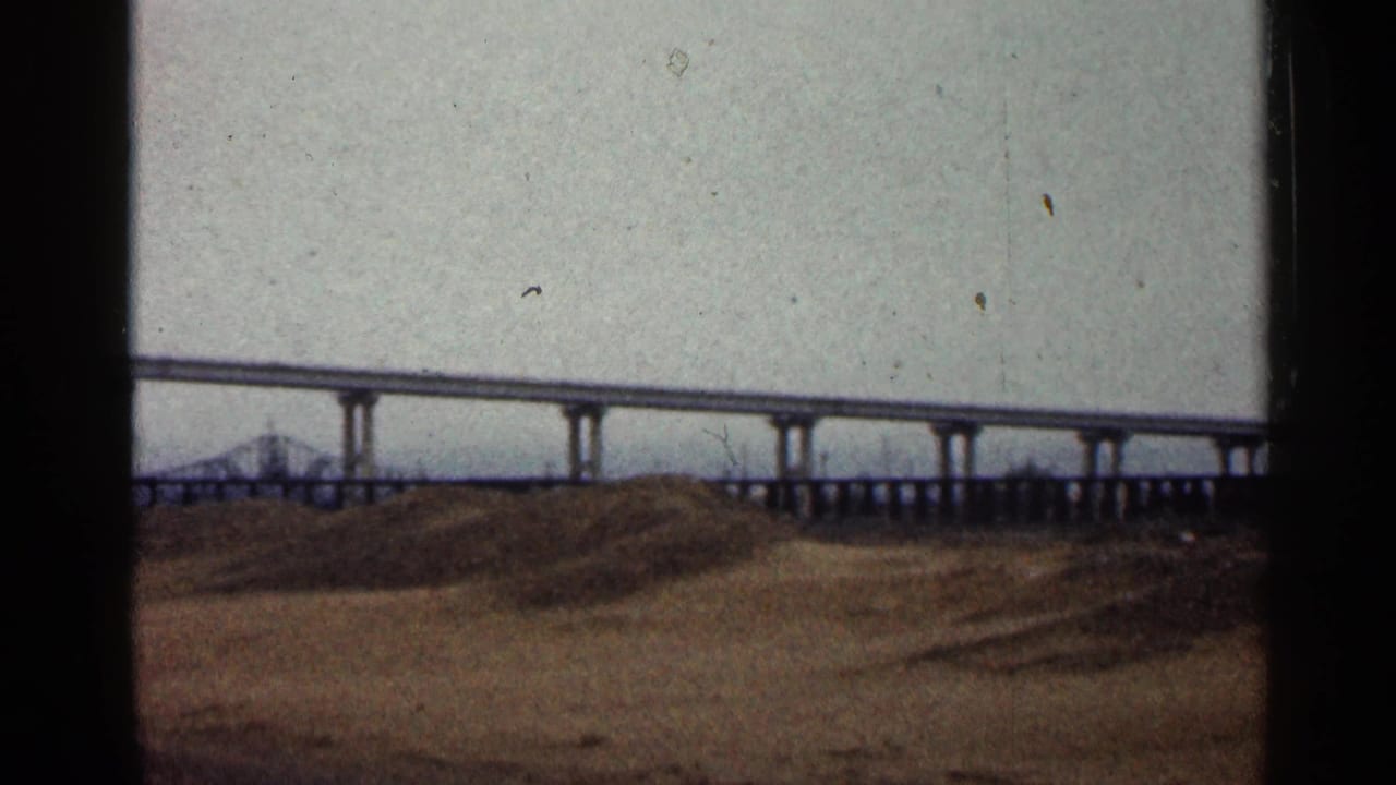 1960 Duluth home-movie frame showing a long concrete overpass above bare brown ground beneath a gray, cloudy sky.