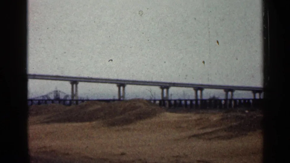 1960 Duluth home-movie frame showing a long concrete overpass above bare brown ground beneath a gray, cloudy sky.