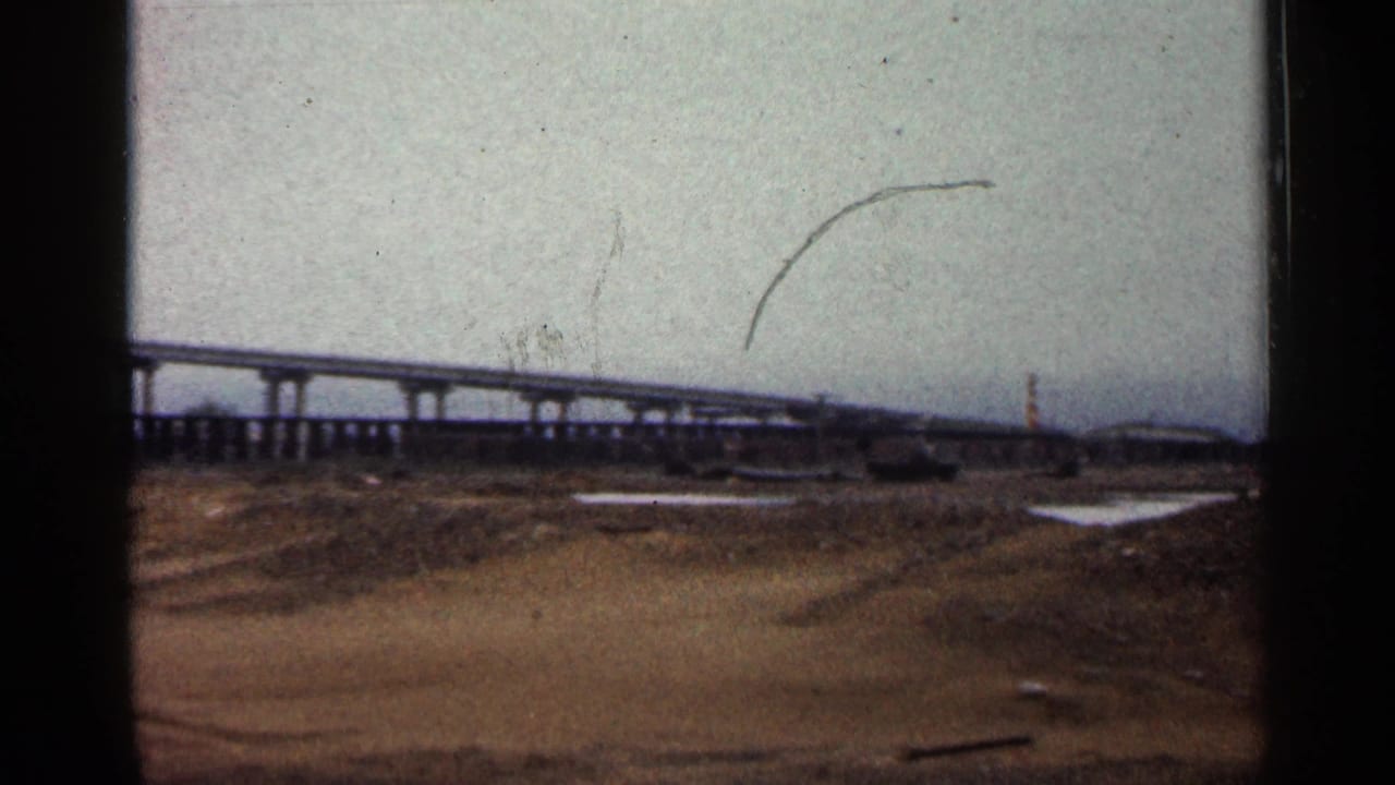 1960 Duluth 8mm frame showing a concrete overpass, exposed brown ground, small puddles, and a cloudy sky.