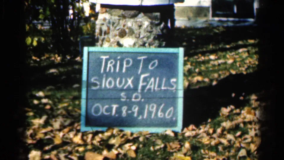 Second view of the 1960 Sioux Falls trip sign, with chalk lettering, leaves, grass, and stonework behind it.
