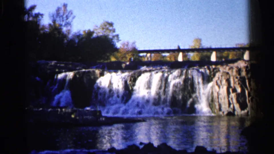 Wide view of a Sioux Falls waterfall with water cascading over rock, trees at the sides, and a bridge crossing above.
