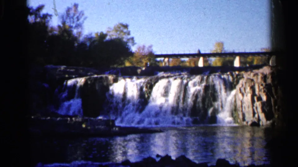 Another 1960 Sioux Falls view shows the waterfall, reflective river water, rocky falls, trees, and bridge in handheld 8mm framing.