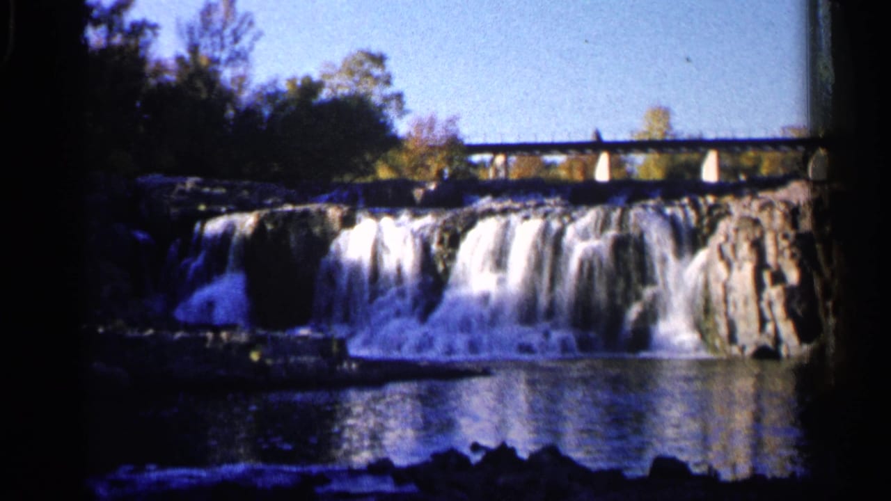 Waterfall and rocky river ledges with a bridge above, framed by trees and film edges.